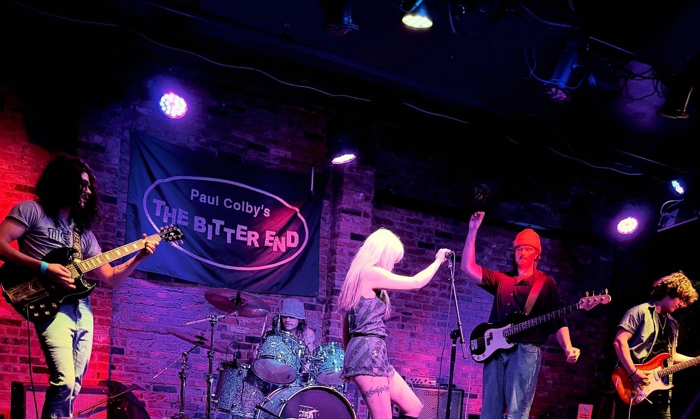 The band on the small stage at Paul Colby's The Bitter End, red and purple light across the brick wall behind them, the singer silhouetted with her back to the camera and one arm raised beside the drum kit, a guitarist on either side and the bassist in a red beanie at the edge of the frame